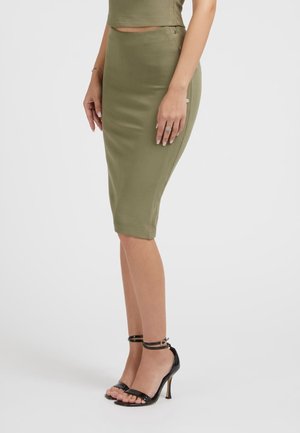 Kvinde iført en olivengrøn knælang pencilskørt, matchende top og sorte højhælede sandaler med ankelrem, stående mod en ensfarvet baggrund.