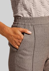 Grijze textured broek met een rekbare tailleband, voorzien van zijzakken en subtiele plooien. Close-up van een hand die in de zak rust.