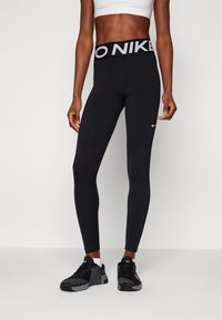 Sportovní osoba oblečená v černých legínách Nike a černých tréninkových botách stojící na bílém pozadí.