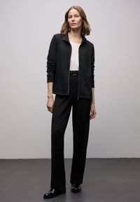 Giacca nera con zip in tessuto morbido, abbinata a una maglietta bianca e pantaloni neri a gamba larga. Texture liscia, design minimalista e stile aderente.