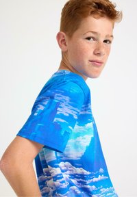 Blå kortærmet T-shirt med et livligt grafik af skyer og byens skyline. Glat tekstur, rund halsudskæring og løs pasform.