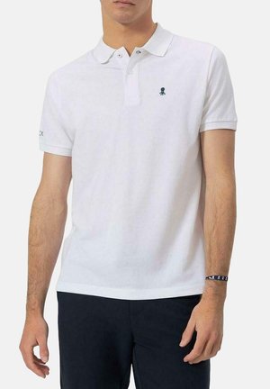 Camisa polo blanca de algodón, con tapeta de tres botones, logo de pulpo bordado y mangas cortas. Combinada con pantalones oscuros.