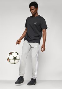 Sort kortærmet t-shirt med et lille logo, grå sweatpants med hvide sidelister og sorte sportssko, der balancerer en mønstret fodbold.