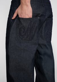 Pantalons en denim foncé avec une coupe slim, featuring des coutures violettes et de grandes lettres brodées sur les poches arrière. Texture lisse et design durable.