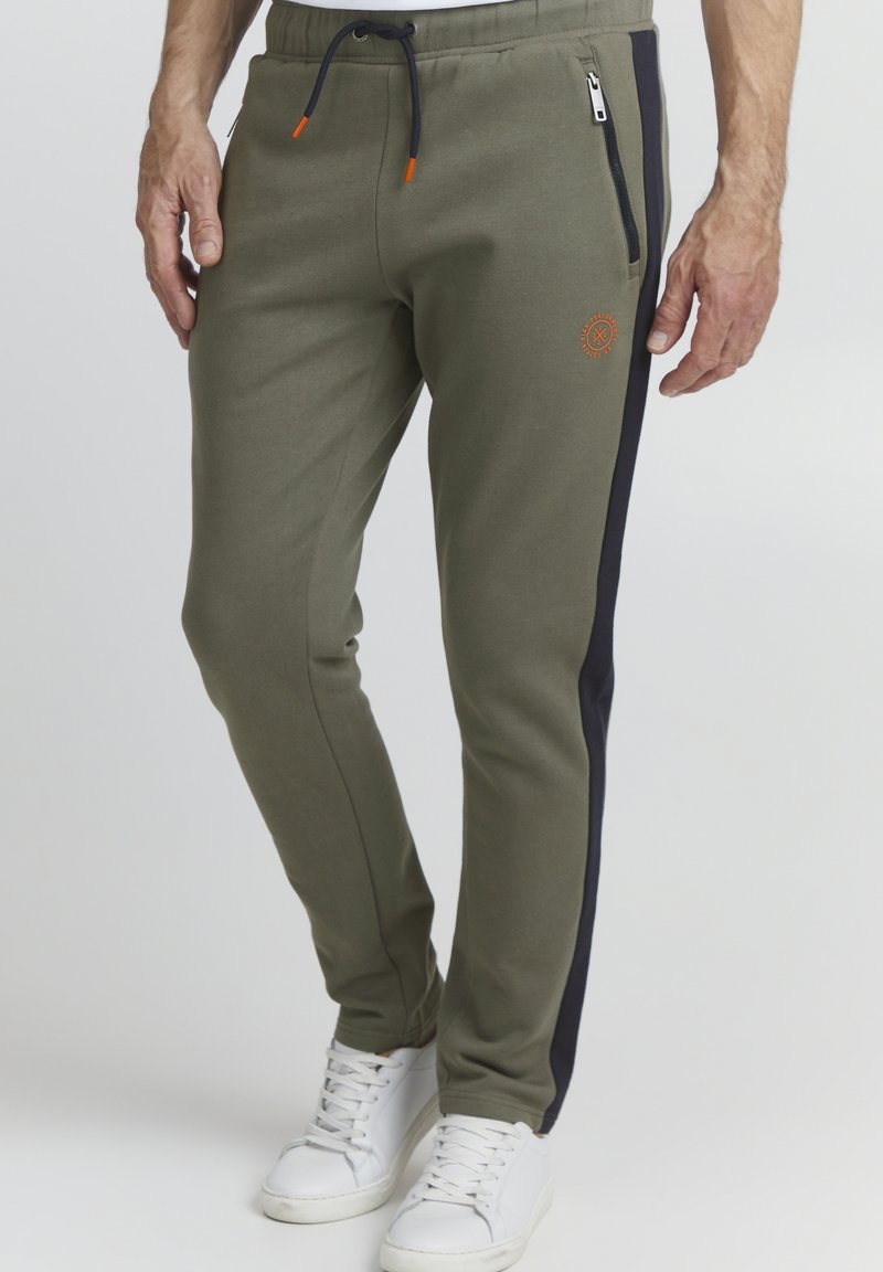 Olive green joggers with navy side stripes, elastic waistband, and drawstring. Features zippered pocket and embroidered logo on leg.