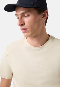Lyse beige t-shirt med korte ærmer, rund halsudskæring og subtilt prægede "BOGNER" logo. Båret med en mørkeblå kasket. Glat tekstur.