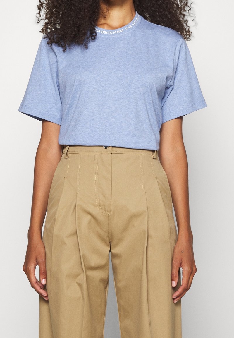 Camiseta de algodón azul claro con mangas cortas, cuello redondo y un sutil detalle de logo alrededor del escote, combinada con pantalones de cintura alta en tono beige.