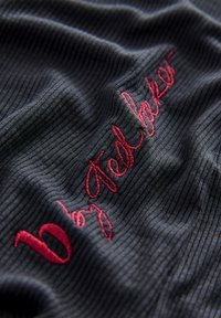 Fekete bordás anyag, melyen piros hímzett szöveg olvasható, miszerint "by Ted Baker." Az anyag puha tapintású, jól látható varrási részletekkel.