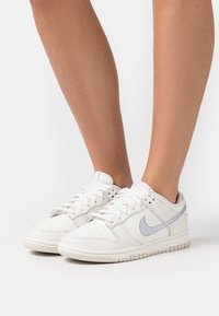 Baskets Nike blanches avec une tige en cuir lisse, des accents gris clair, une boîte à orteils perforée et une semelle en caoutchouc plate. Design lacé pour un ajustement sécurisé.