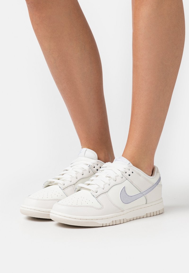 Baskets Nike blanches avec une tige en cuir lisse, des accents gris clair, une boîte à orteils perforée et une semelle en caoutchouc plate. Design lacé pour un ajustement sécurisé.