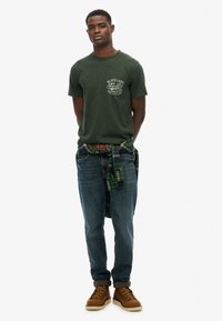 Groene crewneck t-shirt met een grafisch ontwerp op de borst, blauwe jeans en bruine laarzen. Een geruite shirt geknoopt om de taille voegt textuur en kleurcontrast toe.