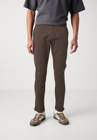 Pantalons marron slim-fit au texture lisse, avec une taille à bouton et des ourlets retroussés, associés à des baskets vertes et marron.