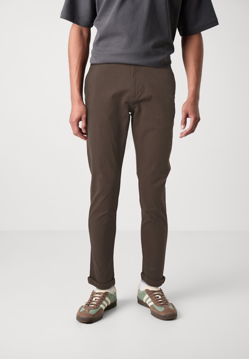Pantalons marron slim-fit au texture lisse, avec une taille à bouton et des ourlets retroussés, associés à des baskets vertes et marron.