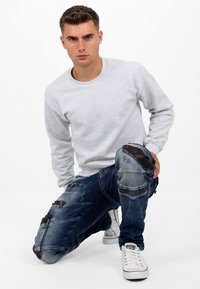 Sweat-shirt gris à manches longues associé à un jean bleu déchiré avec fermetures éclair, patchs aux genoux et accents en cuir noir. Baskets blanches.