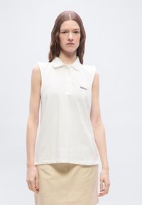 Γυναίκα που φοράει αμάνικο λευκό πόλο με το λογότυπο "Barbour" και μπεζ φούστα, στέκεται μπροστά σε απλό φωτεινό φόντο.