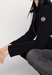 Černá softshellová bunda s nášivkou na levém rukávu, má zipový uzávěr a tvarované rukávy. Nositá přes šedé džíny.