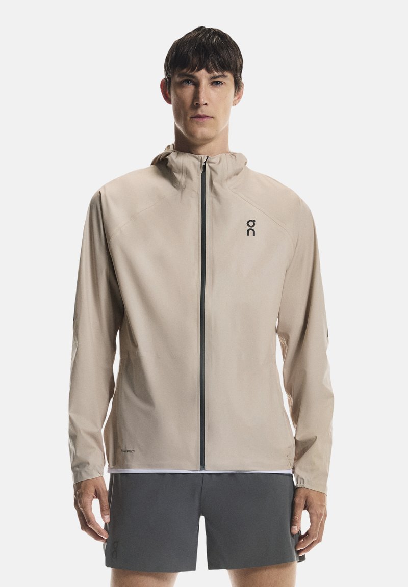 Beżowa lekka kurtka z przednim zamkiem błyskawicznym, kapturem i czarnym logo. Charakteryzuje się opływowym krojem, gładką fakturą i brakiem widocznych wzorów.