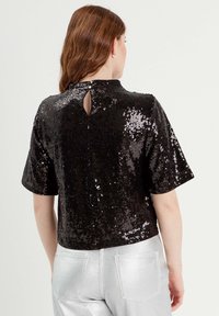 Top negro de lentejuelas con mangas cortas, cuello alto y detalle de abertura en la espalda. Combinado con pantalones metálicos plateados.