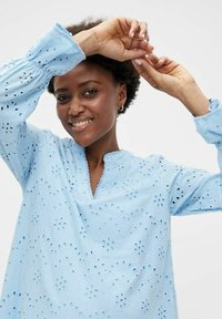 Šviesiai mėlyna siuvinėjimo palaidinė su gėlių iškirpimais, mandarinų apykakle ir ilgomis rankovėmis su surinktomis apykaklėmis. Lygi medvilnės tekstūra.