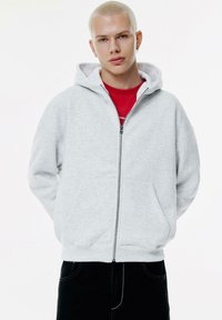 Felpa grigia con zip, cappuccio regolabile, tasche a marsupio e polsini a costine, indossata sopra una maglietta rossa. Tessuto morbido, vestibilità comoda.