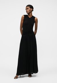 Naine, kes kannab varrukateta musta kortsus bodiisi ja voltidega pikka seelikut, seisab käed selja taga ning vaatab kõrvale.