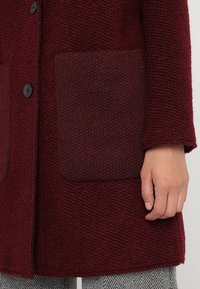 Burgunderfarbener Wollmischmantel mit texturiertem Körper, ausgestattet mit zwei aufgesetzten Taschen und schwarzen Knöpfen, gezeigt mit einer Hand, die den Stoff berührt.