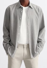 Camicia a quadri bianca e nera con maniche lunghe, indossata sopra una t-shirt bianca. Pantaloni chiari completano l'outfit.