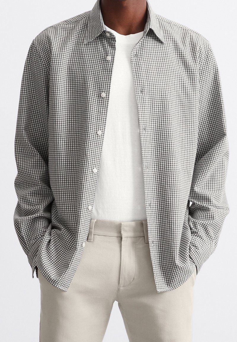 Camicia a quadri bianca e nera con maniche lunghe, indossata sopra una t-shirt bianca. Pantaloni chiari completano l'outfit.