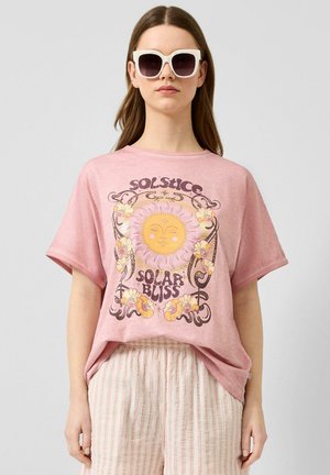 Kobieta nosząca białe okulary przeciwsłoneczne, luźny różowy t-shirt z wzorem słońca i kwiatów oraz napisem, oraz jasne, paskowane spodnie na jednolitym tle.