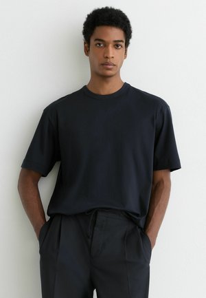 Ung mand med kort, krøllet hår står op ad en hvid væg iført en ensfarvet sort t-shirt og sorte bukser med hænderne i lommerne.