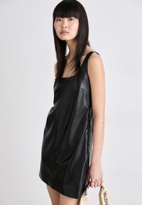 Črna usnjena mini obleka s kvadratnim izrezom, tankimi naramnicami in gladko teksturo. Model drži blagovno znamko z napisom "VALENTINO".