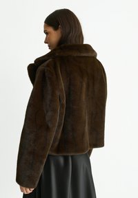 Brown faux fur cropped jacket with a collar, featuring a smooth texture and wide sleeves, paired with a black, flowy garment underneath.