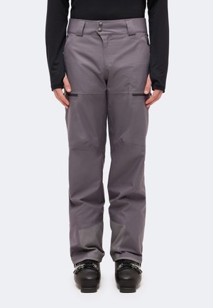 Un mannequin porte un pantalon de randonnée gris aux genoux et aux bas renforcés et se tient debout, chaussé de chaussures de ski noires, sur un fond uni.