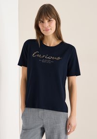 Femme portant un t-shirt bleu marine avec le texte "Curious", assorti à un pantalon à carreaux gris, debout devant un fond beige clair.