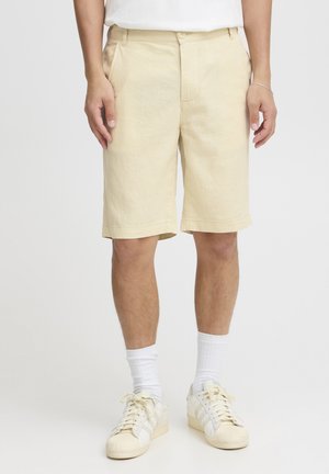 Shorts beiges en tissu texturé, de longueur mi-longue, coupe droite, poches avant et bouton métallique. Associés à des chaussettes blanches et des baskets.