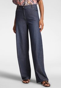Mujer vestida con jeans de mezclilla oscuros de pierna amplia con bolsillos frontales y sandalias marrones, de pie sobre un fondo blanco.