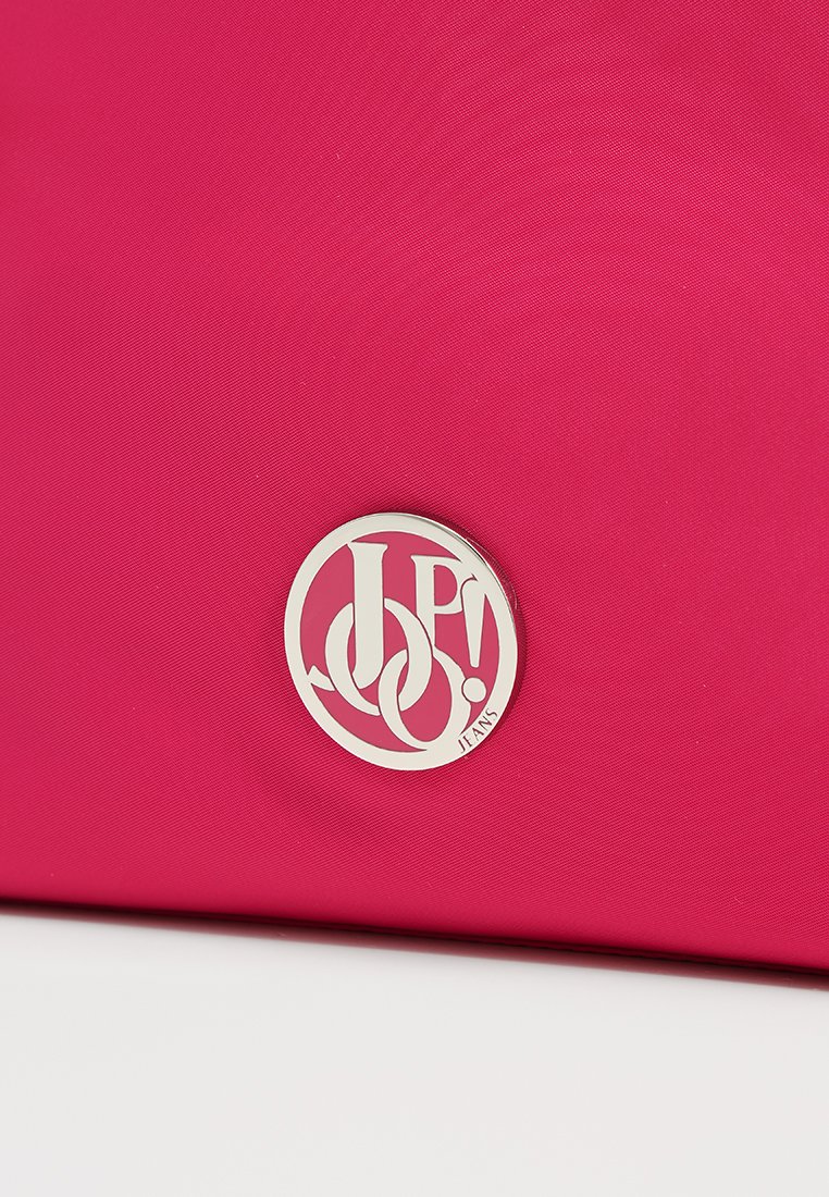 Rosa Stofftasche mit einem auffälligen silbernen, runden Logo in der Mitte, das die ineinander verschlungenen Buchstaben "JP!" und "JEANS" zeigt. Glatte Textur.