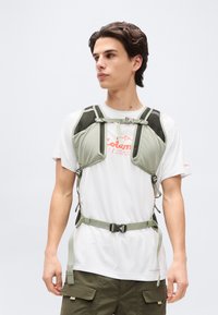 Νεαρός άνδρας με ανοιχτό πράσινο θώρακα πάνω από λευκό γραφικό μπλουζάκι και χακί παντελόνι cargo, κοιτάζει προς τα αριστερά σε απλό λευκό φόντο.