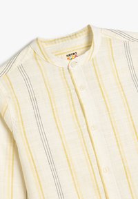 Katoenen kindershirt in crème met gele en zwarte verticale strepen. Het heeft een ronde kraag en drukknoopsluiting met een lichte textuur.