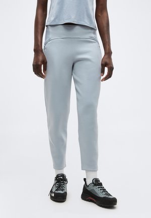 Persoană purtând pantaloni de trening gri deschis, șosete albe și încălțăminte de drumeție gri și neagră, stând în fața unui fundal simplu, deschis.