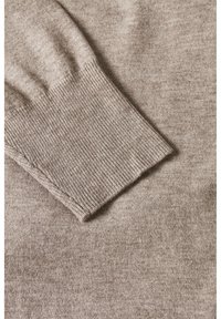 Stickad tröja i beige, med en jämn textur och ribbade ärmslut. Närbilden visar mjuka fibrer och subtila färgvariationer.