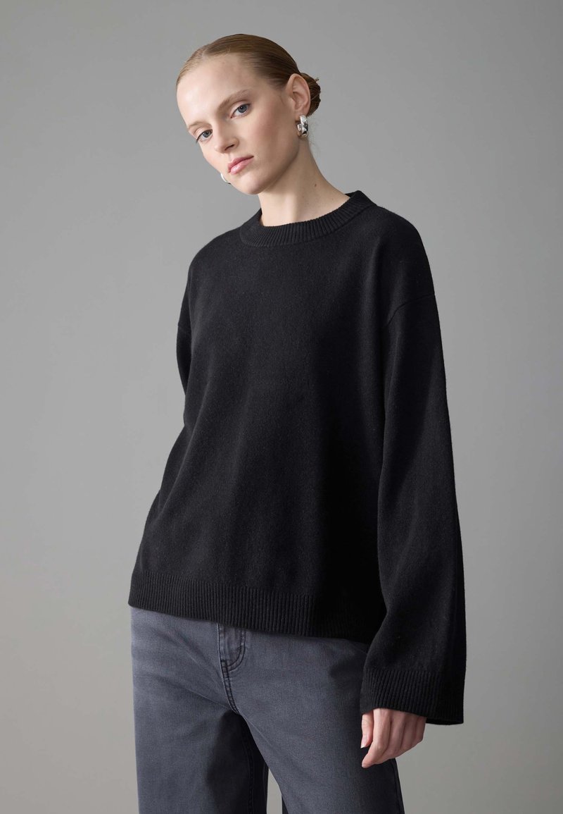 Black knit sweater with a crew neckline, dropped shoulders, and ribbed hem. Worn with gray pants, set against a neutral background.