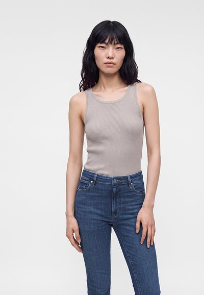 Jeune femme aux cheveux noirs portant un haut gris sans manches et un jean bleu, debout devant un fond clair uni.