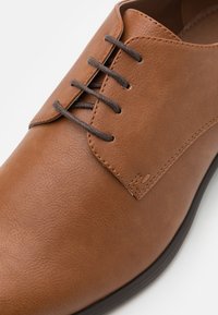 Brown leather shoe with a smooth texture, featuring a lace-up closure, brown laces, and a stitched detailing along the edge.