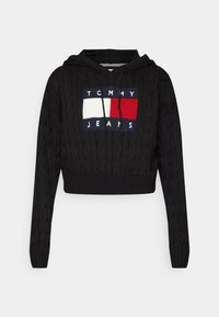 Black cable-knit cropped hoodie with front logo patch in white, red, and navy and "Tommy Jeans" text, featuring drawstrings and ribbed cuffs.