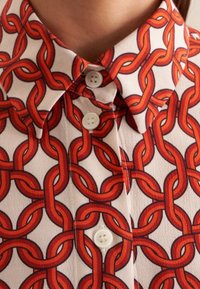 Vit skjorta med djärvt röd sammanflätad kedjeprint, knäppt upp till en spetsig krage.