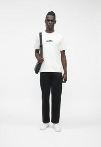 Camiseta blanca de algodón con un logo azul, pantalones de mezclilla negros y zapatillas blancas. La persona lleva un bolso negro de hombro.