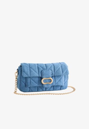 Frau in einem hellgelben schulterfreien Kleid, die eine blaue gesteppte Handtasche mit goldenem Kettengurt hält, steht vor einer schlichten hellen Wand.