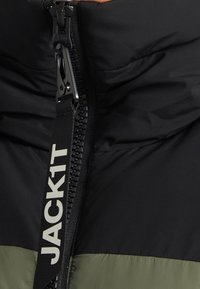 Black and olive green jacket zipper with attached black fabric pull tab labeled "JACKT" in white letters.