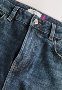 Vysoké rozšírené rifle z tmavomodrého denimu s viditeľným prešívaním. Majú kovové gombíky a predné vrecká, ktoré zdôrazňujú textúrovaný povrch.